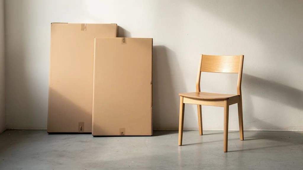 Zwei flache Möbelkartons lehnen an einer Wand neben einem fertig montierten Holzstuhl auf Betonboden