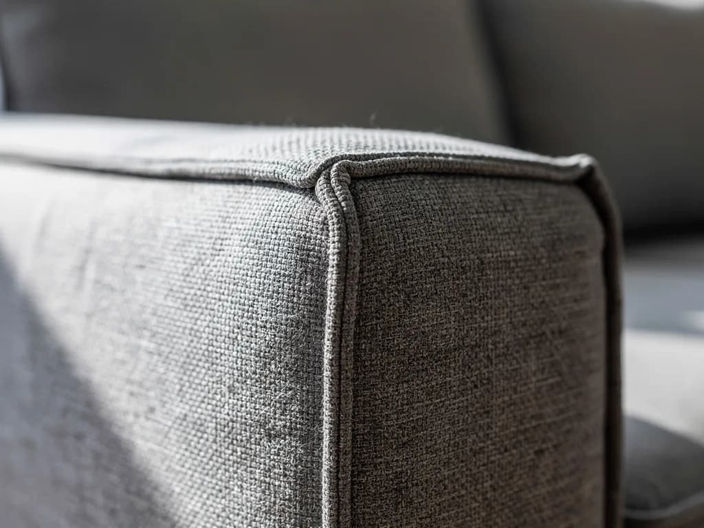 Nahaufnahme der Armlehnenkante eines grauen Sofas mit sichtbarer Kedernaht und grober Gewebestruktur.