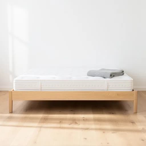Neue weiße Federkernmatratze auf einem hellen Eichen-Bettgestell mit grauer Wolldecke in einem minimalistischen Schlafzimmer.
