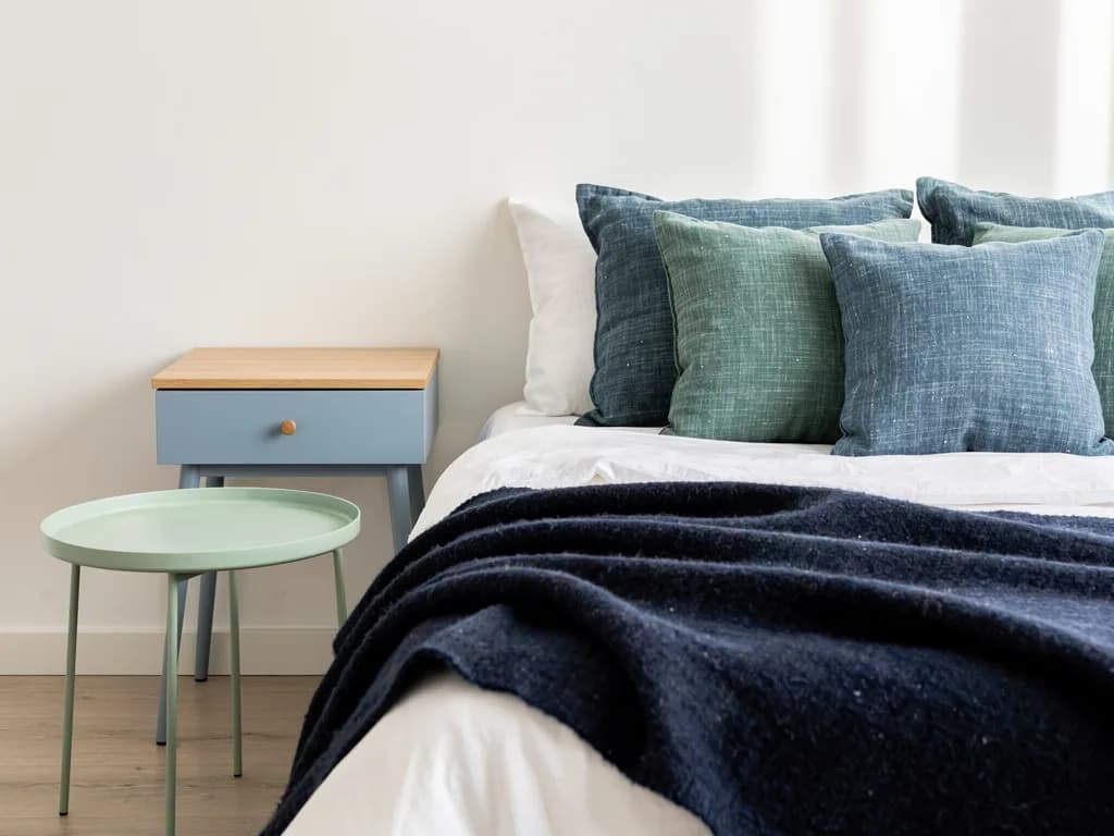 Schlafzimmerecke mit hellblauem Nachtschrank, mintgrünem Beistelltisch und dunkelblauer Wolldecke.