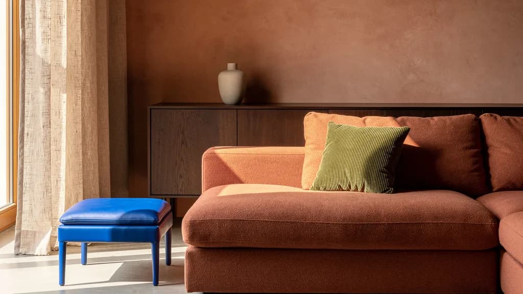Wohnzimmer in Lehmtönen mit terrakottafarbenem Ecksofa, olivgrünem Cordkissen und kobaltblauer Lederbank am Fenster