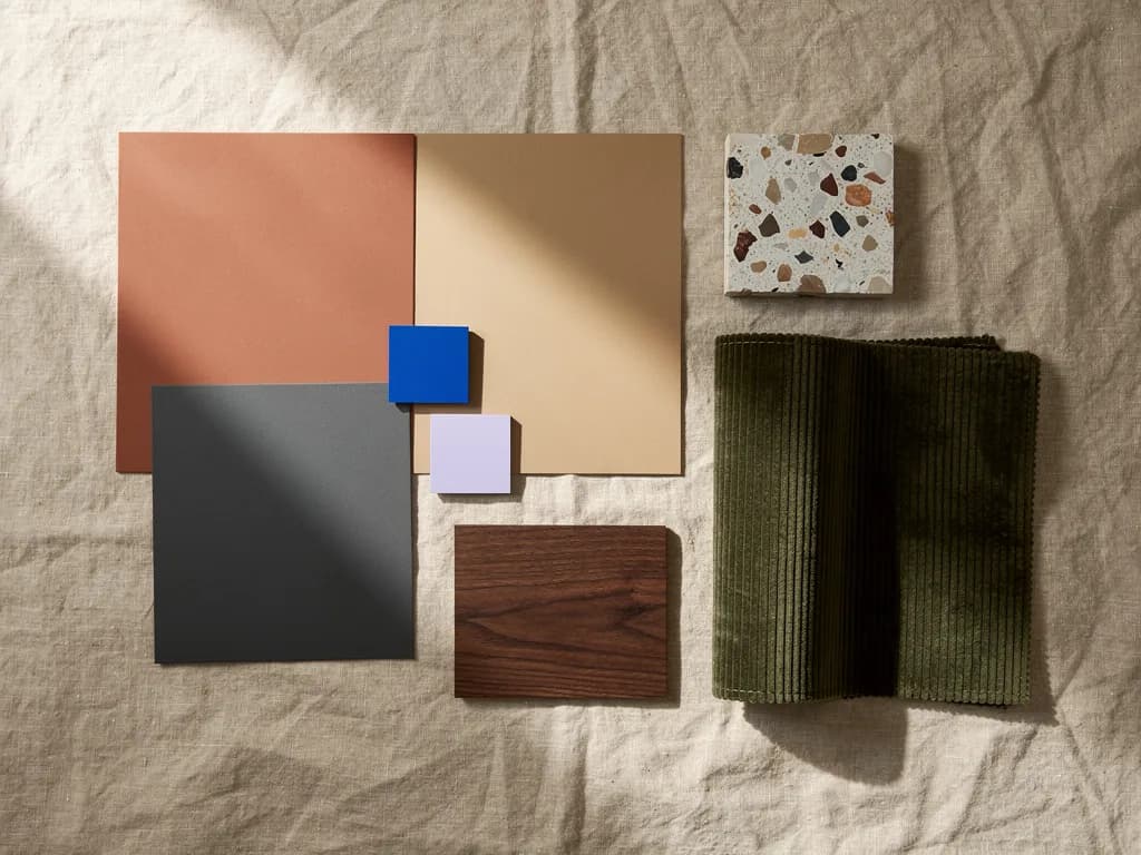 Materialpalette für 2027 auf Leinen mit Lehm, Sand, Ruß, Nussbaum, Terrazzo, olivgrünem Cord und Akzentchips in Kobaltblau und Lila