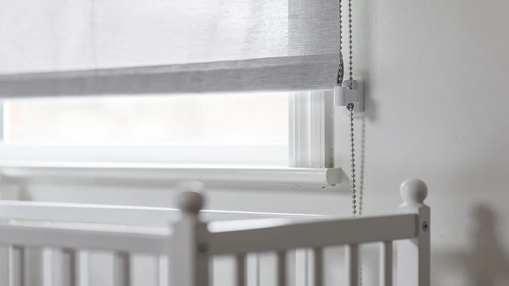 Rollo am Kinderzimmerfenster, dessen Bedienkette in einer weißen Wandspannvorrichtung endet, davor ein weißes Gitterbett.