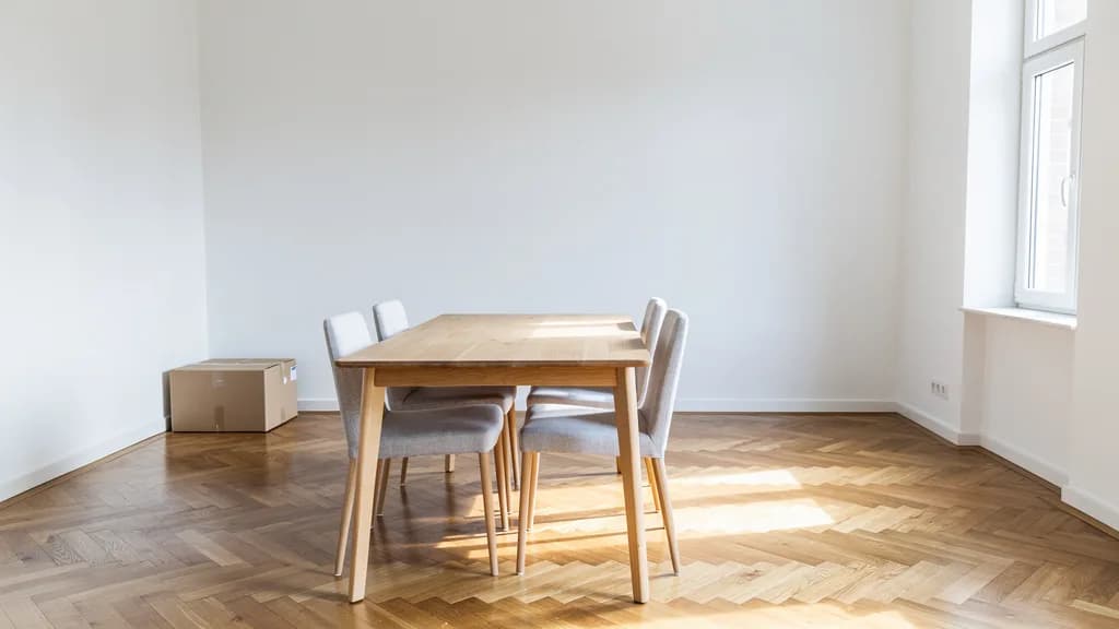 Eichenesstisch mit vier grauen Polsterstühlen in leerer Altbauwohnung mit Fischgrätparkett und einem Umzugskarton.