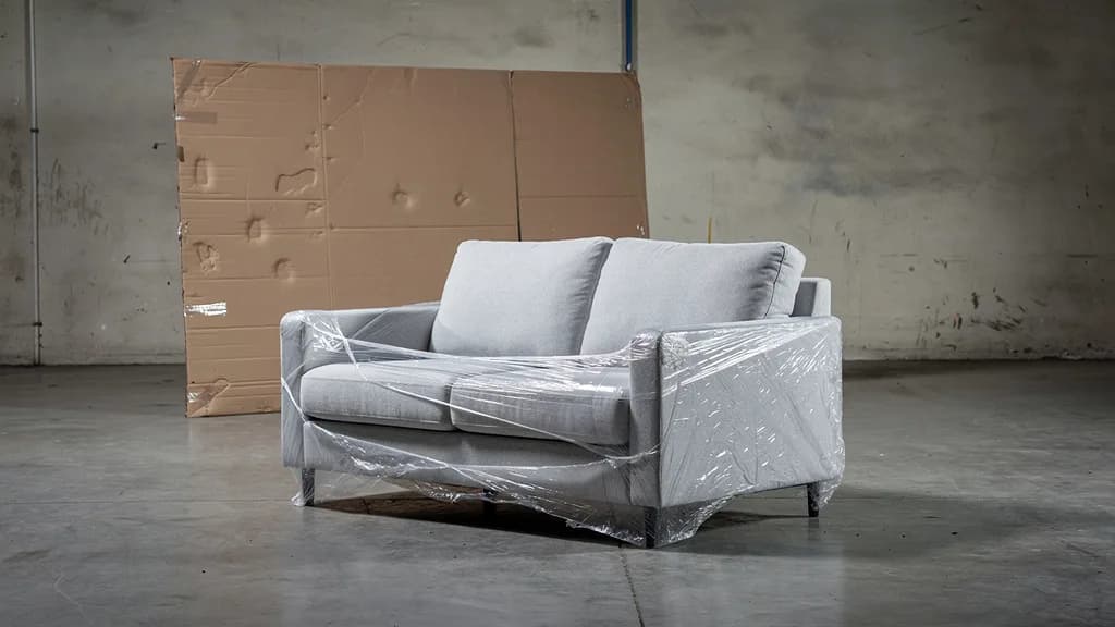 Hellgraues Zweisitzer-Sofa halb in Schutzfolie auf Betonboden, dahinter ein flachgelegter Karton.