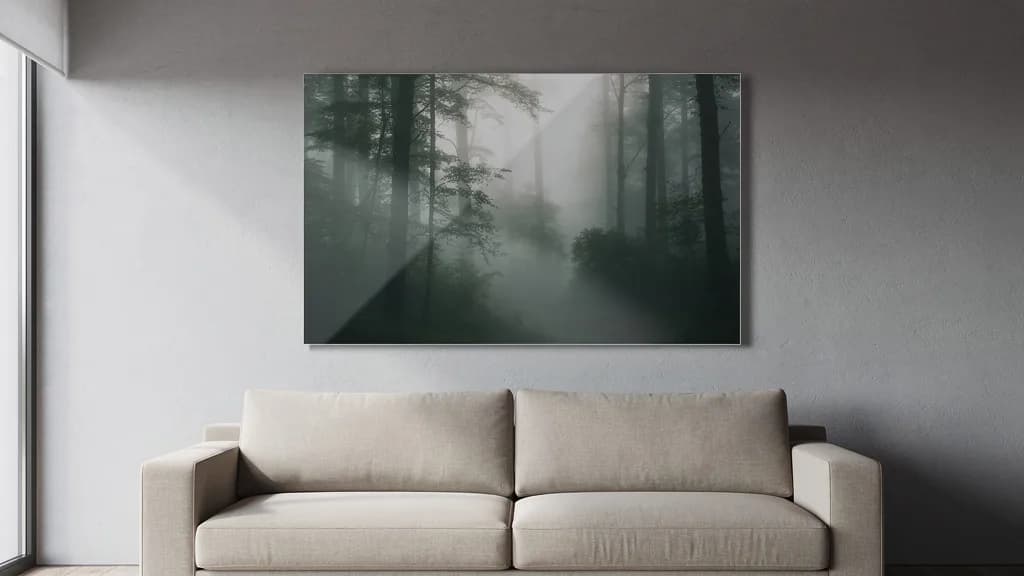 Rahmenloses Glasbild mit Nebelwald-Motiv hängt über einem beigen Stoffsofa an einer grauen Wand.