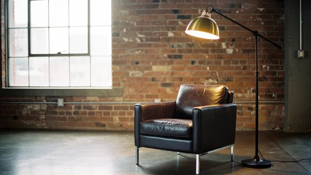 Schwarze Stehlampe mit Gelenkarm und messingfarbenem Schirm neben einem Ledersessel vor einer Ziegelwand in einem Loft
